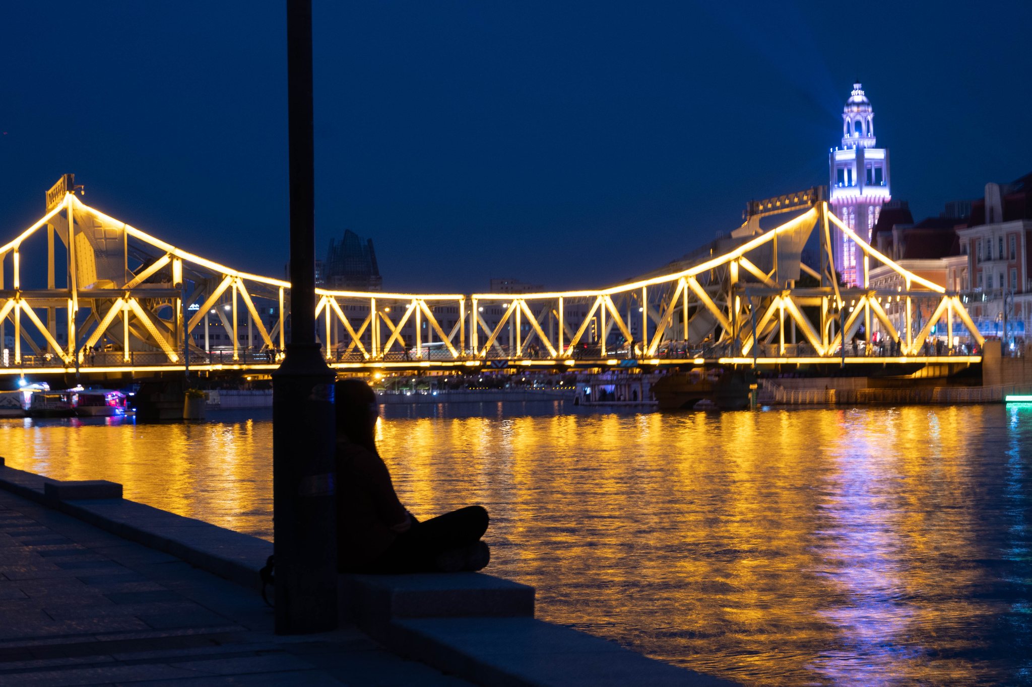 Figure sitting by a lit bridge and river